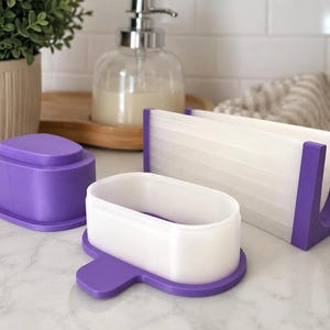 May include: A set of purple and white plastic kitchen tools including a rectangular napkin holder, an oval container with a matching purple lid, and a separate oval base, all featuring a smooth, matte finish and clean, geometric lines.