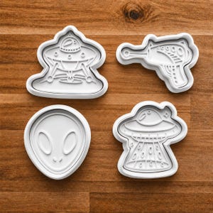 May include: Four white plastic cookie cutters in the shapes of alien-themed designs. The shapes include a ray gun, a classic alien head, and two different flying saucers. The cookie cutters are on a wooden surface.