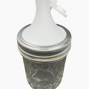 May include: A clear glass jar with a silver metal lid and a white plastic pour spout. The spout has a hinged lid. The jar is designed for dispensing liquids.
