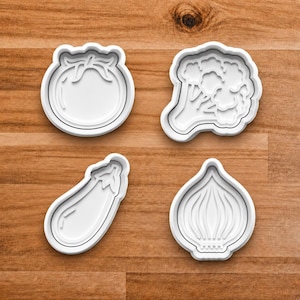May include: Four white plastic cookie cutters in the shapes of vegetables: a tomato, broccoli, an eggplant, and an onion. The cutters are arranged on a wooden surface.