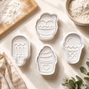 May include: A set of four white plastic cookie cutters featuring ice cream themed designs, including a popsicle, a bowl with scoops, a soft serve swirl with a heart detail, and a classic waffle cone, arranged on a light surface.
