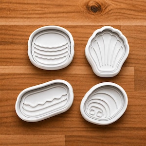 May include: Four white plastic cookie cutters in various shapes: a hamburger, a seashell, a hot dog, and an oyster. The cutters are arranged on a wooden surface, showcasing their detailed designs.
