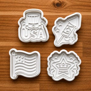 May include: Four white plastic cookie cutters in patriotic shapes: a top hat with "USA" text, a rocket with stars, an American flag, and a star with stripes and stars. The cutters are on a wooden surface.