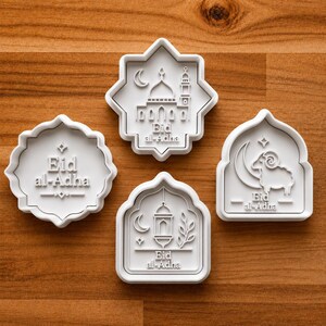 May include: Four white plastic cookie cutters with raised designs for Eid al-Adha. One features a mosque, one a sheep with a crescent moon, one a lantern with a crescent moon, and one features the text Eid al-Adha with decorative accents.