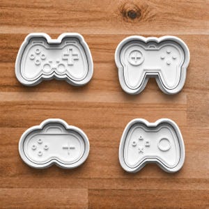 May include: Four white plastic cookie cutters in the shape of video game controllers. Each cutter has a different design, including buttons, directional pads, and joysticks. The cutters are arranged on a wooden surface.