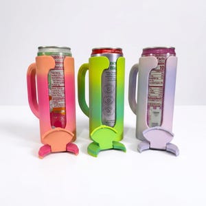 May include: Three colorful can coolers in a row, each holding a beverage can. The coolers are in shades of pink, green, and purple, with a handle and a base. The cans are visible through the cooler's openings.