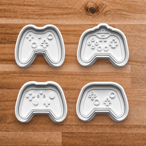 May include: Four white plastic cookie cutters shaped like video game controllers. Each cutter features a distinct button layout including directional pads, action buttons, and joysticks molded into the center, designed for creating shaped cookies.
