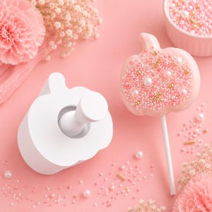May include: A white pumpkin-shaped cookie cutter and a matching pumpkin-shaped cookie on a stick, decorated with pink and white sprinkles, gold rods, and white pearl beads, surrounded by pink flowers and loose sprinkles on a pink surface.