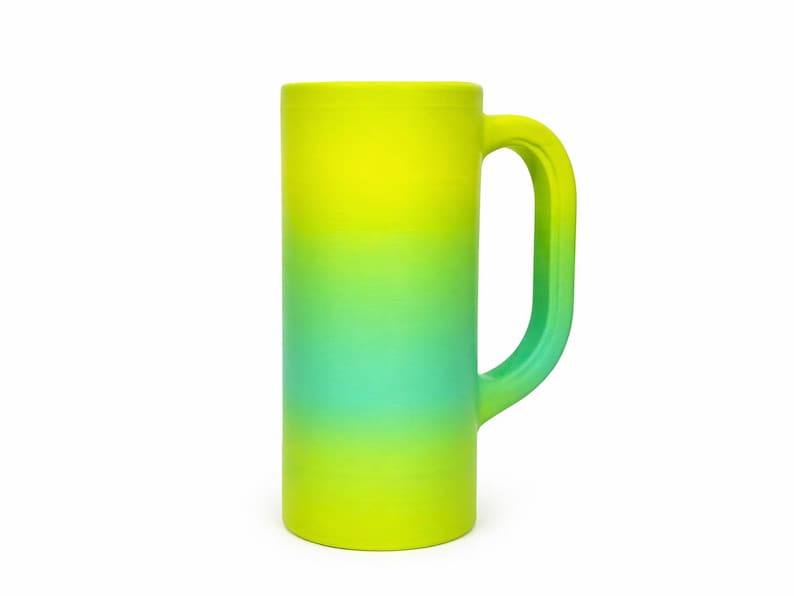 May include: A tall, cylindrical mug with a large handle. The mug features a gradient design, transitioning from yellow at the top to green in the middle and back to yellow at the bottom. The mug is made of a solid material and is suitable for beverages.