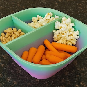 May include: A divided snack container filled with peanuts, popcorn, and baby carrots. The container is a gradient of green and purple. The snacks are arranged in separate compartments.