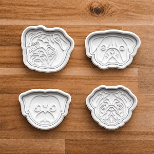 May include: Four white plastic pug-shaped cookie cutters. Each cutter features a different pug face design, with detailed facial features. The cutters are arranged on a wooden surface.