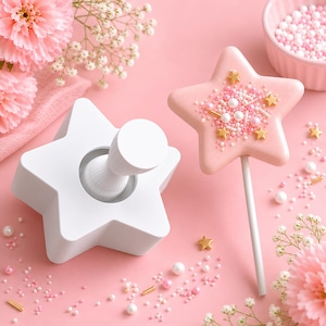 May include: A white star-shaped cookie cutter with a central handle and a matching star-shaped cookie on a stick, decorated with pink icing, white pearl sprinkles, and small gold star-shaped sprinkles, arranged on a solid pink surface.