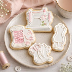May include: Four sugar cookies decorated with royal icing in sewing-themed shapes: a sewing machine, a spool of thread with buttons, a pair of scissors with a measuring tape, and a dress form. The icing is white with accents of soft pink and green.