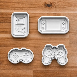 May include: Four white plastic cookie cutters in the shape of retro video game controllers and consoles. The set includes a handheld console, a classic controller, and two other controller designs.