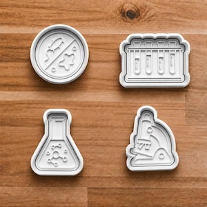 May include: Four white plastic cookie cutters in science-themed shapes: a petri dish with a no-entry symbol and bacteria, a rack of test tubes, a flask with a molecular structure, and a microscope. The cutters are on a wooden surface.