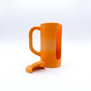 May include: An orange mug with a handle and a large cutout on the side. A detached orange lid sits in front of the mug. The mug is made of a solid material and has a cylindrical shape.
