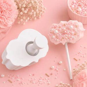 May include: A white plastic scalloped-edge cookie cutter with a central handle and a matching cake pop decorated with pink icing, white and pink sugar pearls, and gold sprinkles on a white stick, surrounded by scattered edible sugar beads.