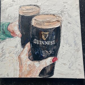 May include: An oil painting on canvas featuring two dark stout beers in pint glasses with thick creamy heads. The glasses are held by hands, one with red nail polish, against a textured white background. The Guinness logo is visible on the glass.