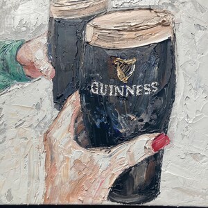 May include: An oil painting with thick impasto texture depicting two dark glasses of stout beer. The glasses feature the gold harp logo and the white text GUINNESS. One glass is held by a hand with red nail polish, and the other by a sleeve.