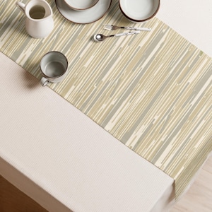 May include: A rectangular table runner featuring a vertical striped pattern in shades of beige, cream, and muted gray. The runner is placed on a white textured tablecloth, accompanied by a white pitcher, two white plates, a cup, and a silver spoon.