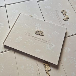 May include: A beige photo album with a floral pattern and the text "Mes souvenirs d'&eacute;cole". A wooden name tag that says "Ella" is attached. Additional albums are visible in the background.