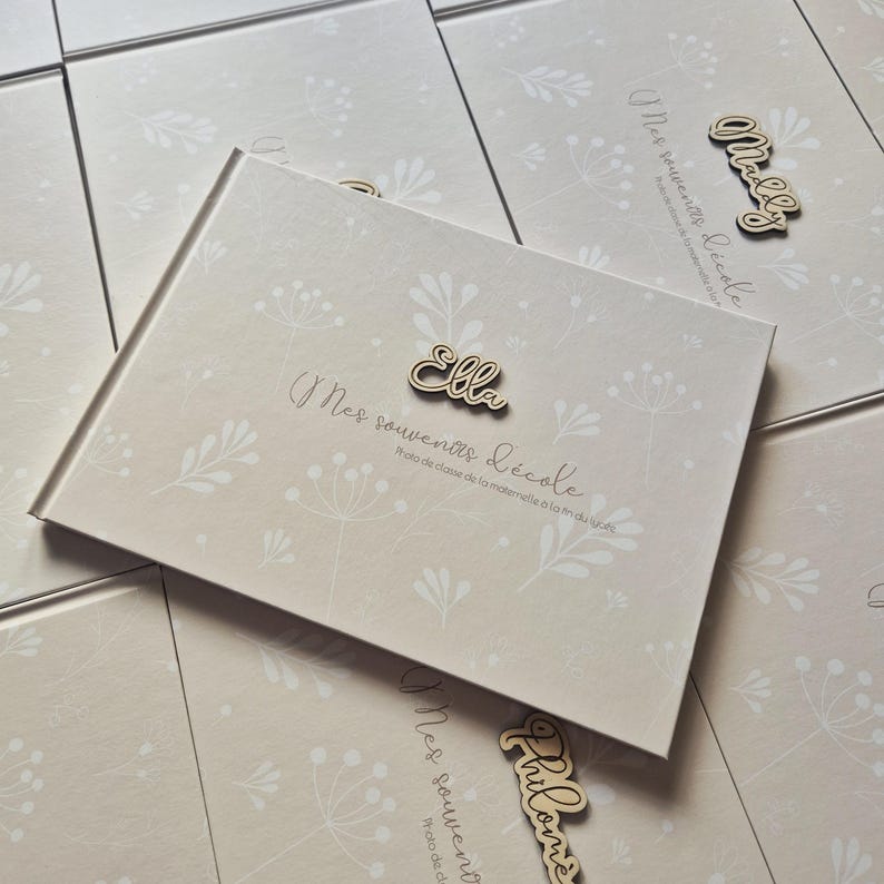 May include: A beige photo album with a floral pattern and the text "Mes souvenirs d'&eacute;cole". A wooden name tag that says "Ella" is attached. Additional albums are visible in the background.