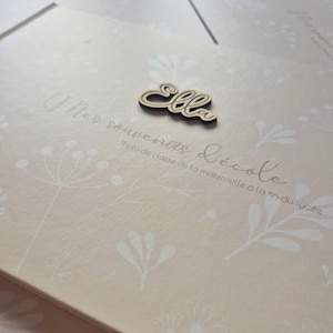 May include: A light pink photo album with a floral design and the name "Ella" in wooden script. The album has the text "Mes souvenirs d'&eacute;cole" and "Photo de classe de la maternelle &agrave; la fin du lyc&eacute;e" printed on the cover.
