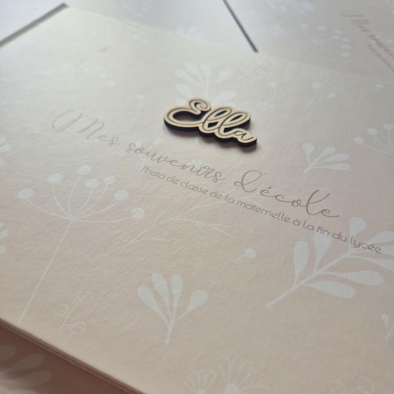 May include: A light pink photo album with a floral design and the name "Ella" in wooden script. The album has the text "Mes souvenirs d'&eacute;cole" and "Photo de classe de la maternelle &agrave; la fin du lyc&eacute;e" printed on the cover.