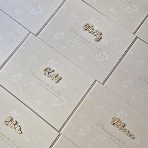 May include: A collection of light beige notebooks with floral patterns and wooden name cutouts. The text "Mes souvenirs d'&eacute;cole" is printed on each notebook. Names like "Maddy", "Kalel", and "Celeste" are visible.
