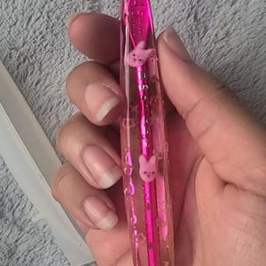 May include: A pink crochet hook with a clear handle, decorated with small pink bunny figures. The hook has a metallic finish and is designed for crafting. The handle is filled with a clear liquid and the hook is held in a hand.