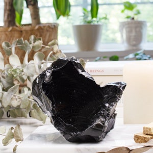 Black obsidian crystal takes the spotlight on a sleek marble backdrop adorned with dried flowers and lush plants, creating a captivating contrast between the crystal's intensity and the soothing beauty of nature.