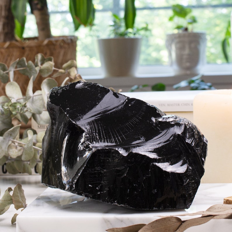 Black obsidian crystal takes the spotlight on a sleek marble backdrop adorned with dried flowers and lush plants, creating a captivating contrast between the crystal's intensity and the soothing beauty of nature. Feng Shui and Negative Energy.