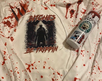 Pode incluir: Casaco de moletom branco com um design de tinta vermelha salpicada. O moletom tem uma estampa gráfica de uma figura mascarada com o texto "Bitches Be Trippin".