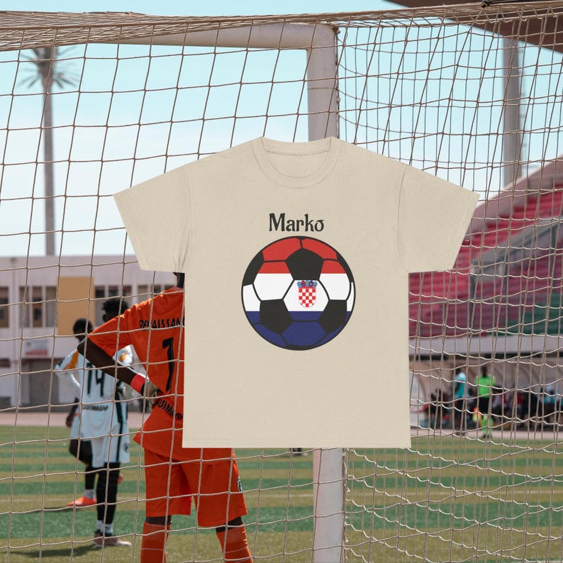 Puede incluir: Una camiseta beige con un dise&ntilde;o de bal&oacute;n de f&uacute;tbol con los colores de la bandera croata y el nombre "Marko" encima. La camiseta se muestra frente a una red de porter&iacute;a de f&uacute;tbol, con jugadores en el fondo.