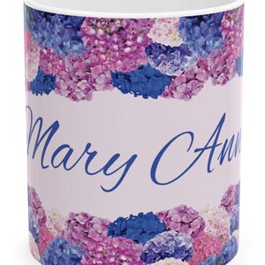 Op de afbeelding: Witte keramische mok met een bloemenontwerp met roze, blauwe en paarse hortensia's. De mok heeft de tekst "Mary Ann" in blauwe cursieve letters.