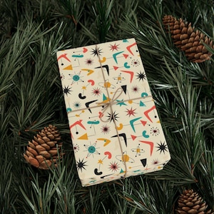 May include: A wrapped gift with a retro atomic pattern in red, yellow, teal, and black on an off-white background. The gift is tied with twine and rests on a bed of pine needles with pine cones.