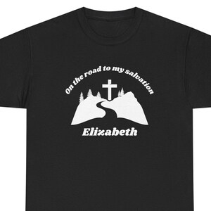 May include: Black t-shirt with a white graphic. The graphic shows a cross above a road winding through mountains, with the text "On the road to my salvation" and the name "Elizabeth".