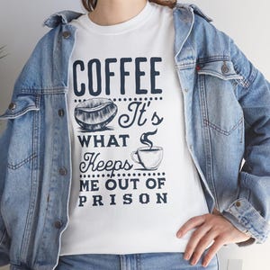 May include: White t-shirt with a navy blue graphic that reads "COFFEE It's WHAT Keeps ME OUT OF PRISON." The design includes a coffee bean and a coffee cup. The shirt is worn with a light blue denim jacket.