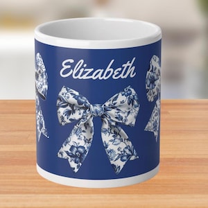 Peut inclure: Une tasse en céramique blanche avec un fond bleu et un nœud blanc avec un motif floral bleu. La tasse porte le nom "Elizabeth" imprimé dessus.