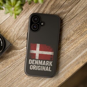 Könnte beinhalten: Schwarze Handyhülle mit der dänischen Flagge in Rot und Weiß im Used-Look. Der Text "DENMARK ORIGINAL" ist darunter aufgedruckt. Die Hülle hat einen Kameraausschnitt und liegt auf einer Holzoberfläche.