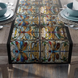 Puede incluir: Camino de mesa rectangular con un diseño de libélulas de vidrieras. El patrón repetido presenta libélulas coloridas en tonos de azul, naranja, amarillo y verde, con un borde negro. El camino de mesa está sobre una mesa de madera.