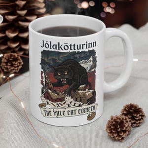 Puede incluir: Una taza de cerámica blanca con una ilustración gráfica de un gran gato negro sobre un pueblo nevado. El texto Jólakötturinn aparece en la parte superior y el texto THE YULE CAT COMETH está impreso en una pancarta en la parte inferior.
