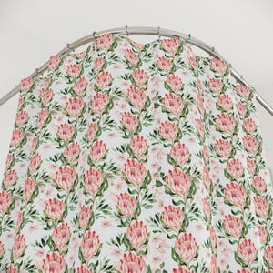 May include: A white shower curtain featuring a repeating pattern of pink protea flowers, green leaves, and smaller pink blossoms. The curtain hangs from a curved metal rail.