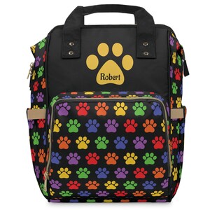 May include: Black backpack with a colorful paw print pattern. The front pocket and sides feature a repeating pattern of rainbow-colored paw prints. The main compartment has a large yellow paw print with the name "Robert" in the center.