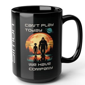 May include: Black ceramic mug with a wraparound graphic featuring an alien and Bigfoot walking in front of a sunset. The text reads "Can't Play Today, We Have Company". The mug has a large handle and the word "Edition" printed vertically on the side.