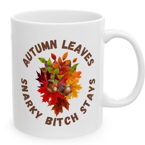 以下が含まれることがあります： 白いセラミックマグカップ、ハンドル付き、秋をテーマにしたデザイン。デザインには、カラフルな秋の葉、ドングリ、そして「AUTUMN LEAVES SNARKY BITCH STAYS」のテキストが円形に配置されています。温かい飲み物にも冷たい飲み物にも適しています。