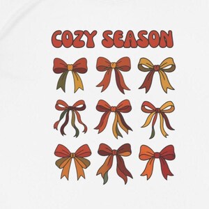 Könnte beinhalten: Weißes T-Shirt mit dem Text "COZY SEASON" in Orange und Braun über neun bunten Schleifenillustrationen. Die Schleifen sind in Rot-, Orange-, Gelb- und Grüntönen gehalten.