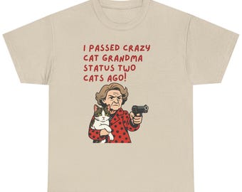 May include: Beige t-shirt with the text "I PASSED CRAZY CAT GRANDMA STATUS TWO CATS AGO!". The graphic shows a woman holding a cat and a gun. The woman is wearing a red jacket with black polka dots.
