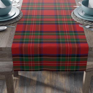 May include: A rectangular table runner featuring a classic tartan plaid pattern in vibrant red, forest green, navy blue, and thin yellow lines. The fabric has a woven texture and is draped across a wooden table surface between two place settings.
