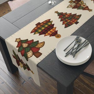 May include: A cream-colored table runner featuring appliqued Christmas tree designs in plaid patterns of red, green, and gold. The runner is placed on a dark wooden table, with white plates and silverware.